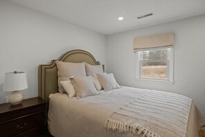 A bedroom with a bed, bedside table, lamp, window with blinds, and a view of the outdoors.