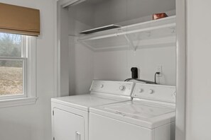 A laundry room with a washer and dryer, a window with a blind, and a shelf with a box.
