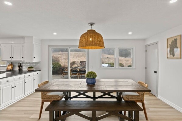 A spacious kitchen with white cabinets, a wooden dining table, wicker chairs, and a large window overlooking a landscape.