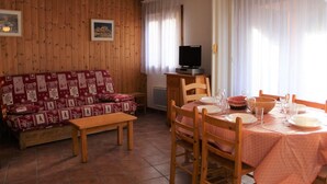 Un salon chaleureux avec un mur en bois, un canapé rouge à motifs, une table basse en bois et un coin repas avec une table dressée pour quatre personnes.