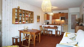 Ett kompakt bostadsutrymme med en bokhylla i trä, ett matbord med stolar, en liten köksdel och en soffa.