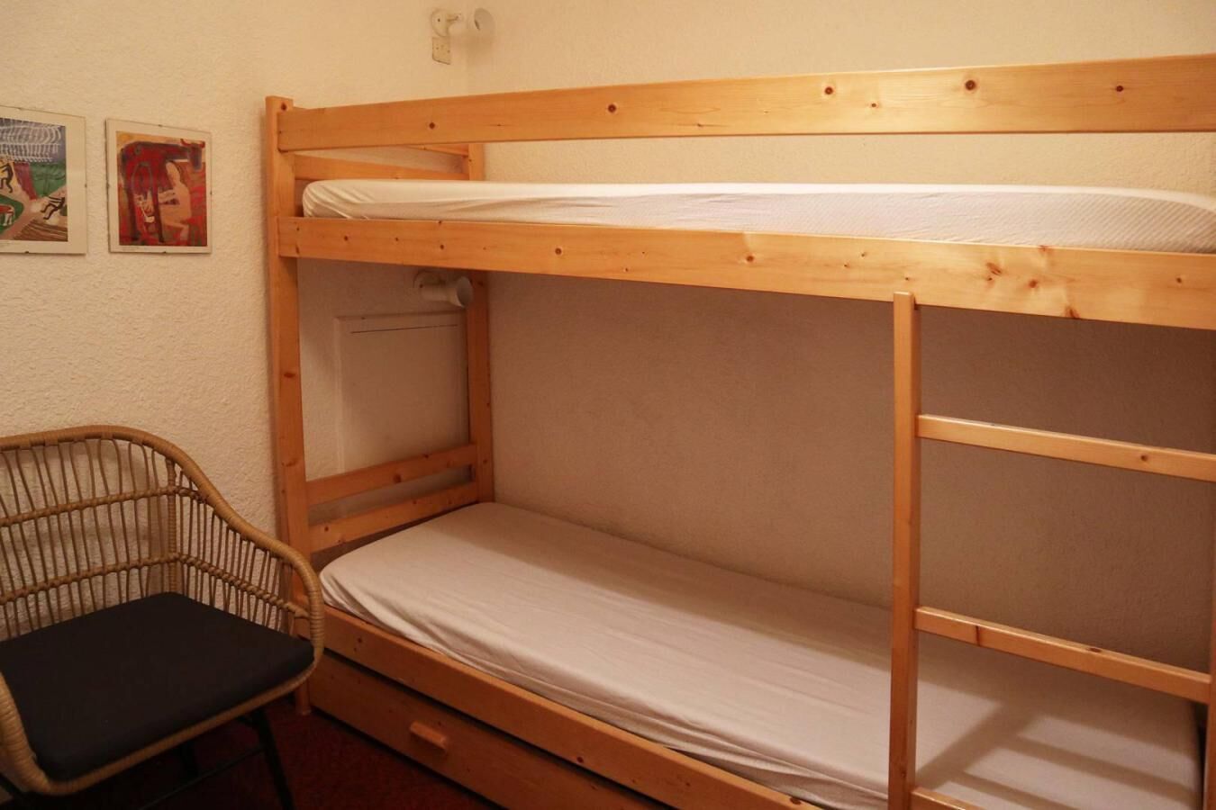 A bunk bed with a wooden frame, a single bed below, and a wicker chair with a cushion.