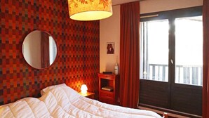Ett hotellrum med en säng, en rund spegel, en vägg med ett rutmönster, ett nattduksbord med en lampa, ett litet bord med en vas och ett fönster med gardiner.
