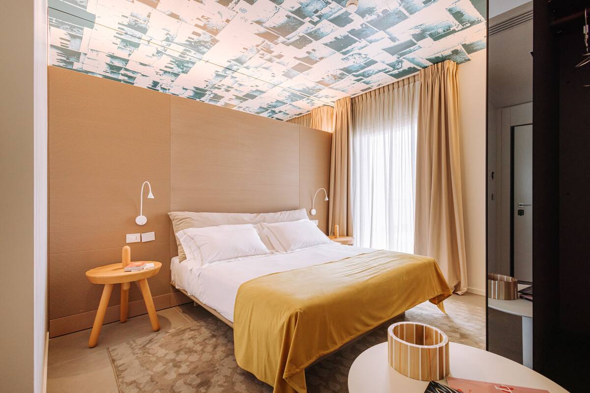 Ein Hotelzimmer mit Bett, Nachttisch, Lampe, einem kleinen Hocker, einem runden Tisch und einem an der Wand befestigten Regal.