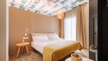 A hotel room with a bed, a bedside table, a lamp, a small stool, a round table, and a wall-mounted shelf.