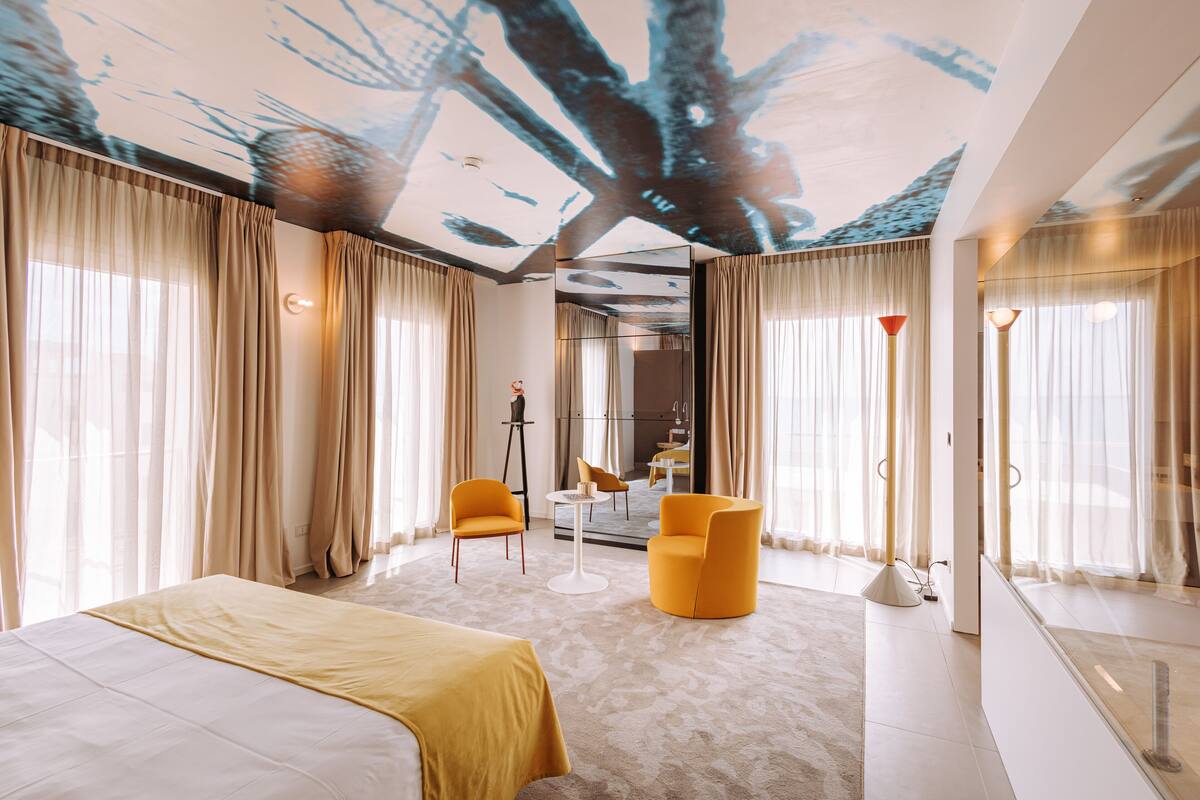 Ein modernes Hotelzimmer mit einem großen Bett, zwei gelben Sesseln, einem kleinen Tisch und einer Deckenprojektion.
