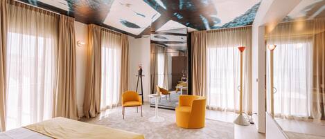 A modern hotel room with a large bed, two yellow armchairs, a small table, and a ceiling with a tree mural.
