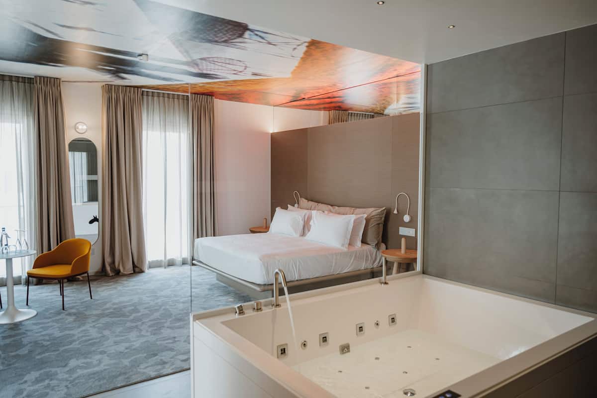 Une chambre d’hôtel moderne dotée d’un grand lit, d’une salle de bain séparée avec une baignoire, d’un coin repas avec une table et des chaises, et d’une grande fenêtre avec des rideaux.