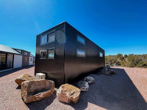 A modern, black metal building with multiple windows, surrounded by rocks and gravel, set against a clear blue sky.