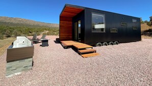 A compact, modern cabin with a wooden exterior and a large window, situated on a gravel surface with a clear sky and distant hills. - The Phoenix (Cañon City)