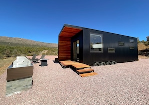 A compact, modern cabin with a wooden exterior and a large window, situated on a gravel surface with a clear sky and distant hills. - The Phoenix (Cañon City)