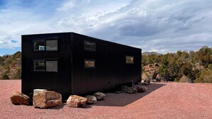 A modern, black, modular building with multiple windows, situated on a gravel surface with large rocks around it, set against a backdrop of trees and distant mountains. - The Phoenix (Cañon City)