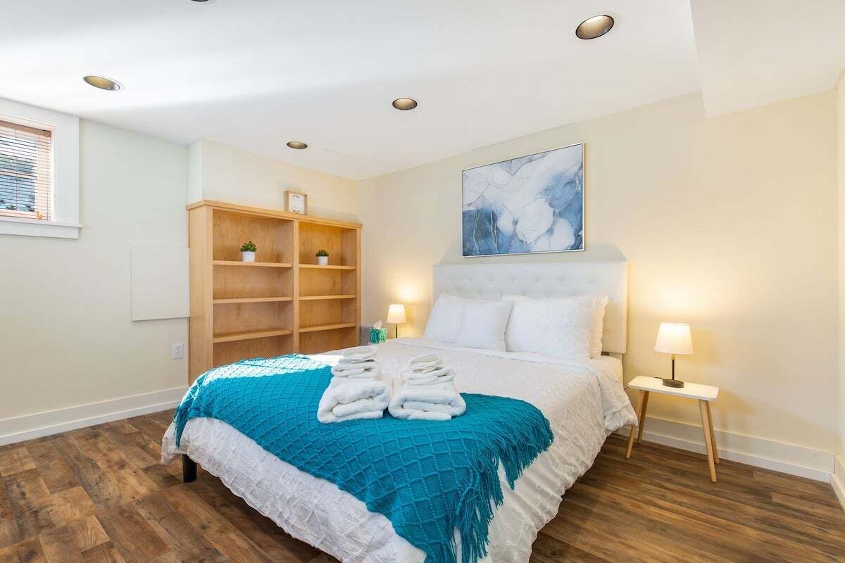 A neatly arranged bedroom with a bed, bedside lamps, a wooden shelf unit, and a painting on the wall.