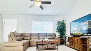A living room with a sectional sofa, a coffee table with baskets, a wooden TV stand, and a ceiling fan. - Comfy and Cozy 4 bedroom home with garage (Ocala)