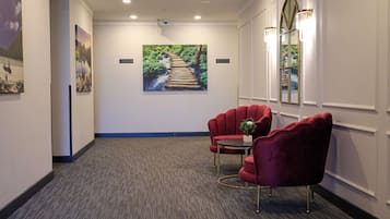 A hotel lobby with a carpeted floor, red upholstered chairs, a large framed picture of a bridge, and a mirror with lights.