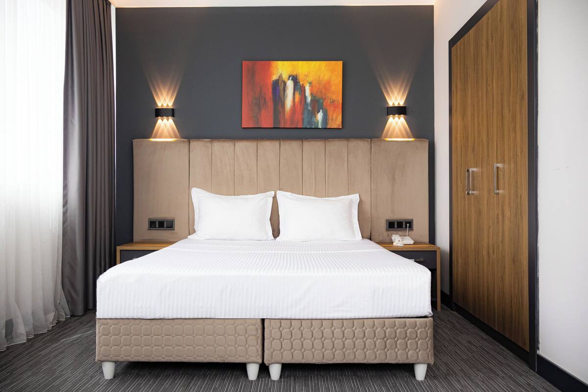 A hotel room with a large bed, two bedside tables, a nightstand, a wooden wardrobe, and a painting on the wall.