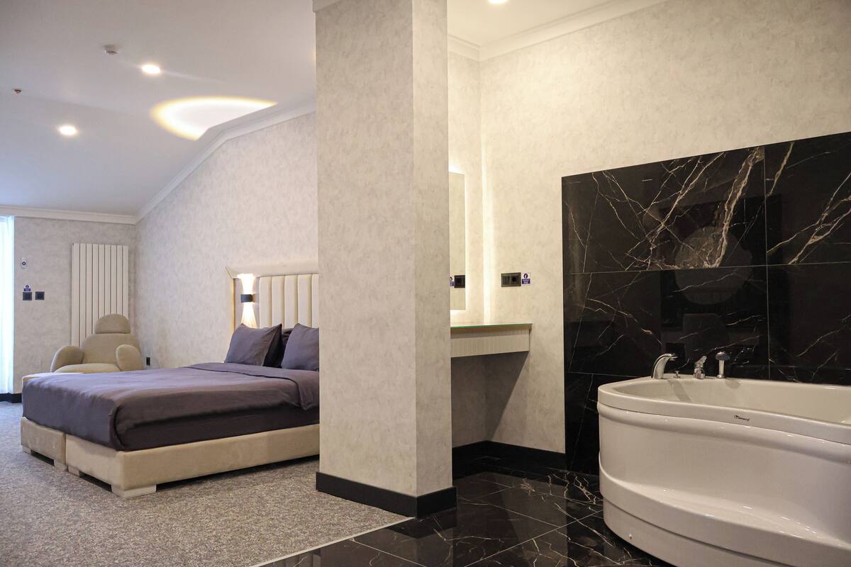 A modern bedroom with a large bed, a sitting area, and a bathroom with a bathtub and marble wall.