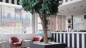 A modern hotel lobby with a reception desk, two pink armchairs, a large artificial tree, and a view of the cityscape through large windows. - HOME PLUS HOTEL (Istanbul)