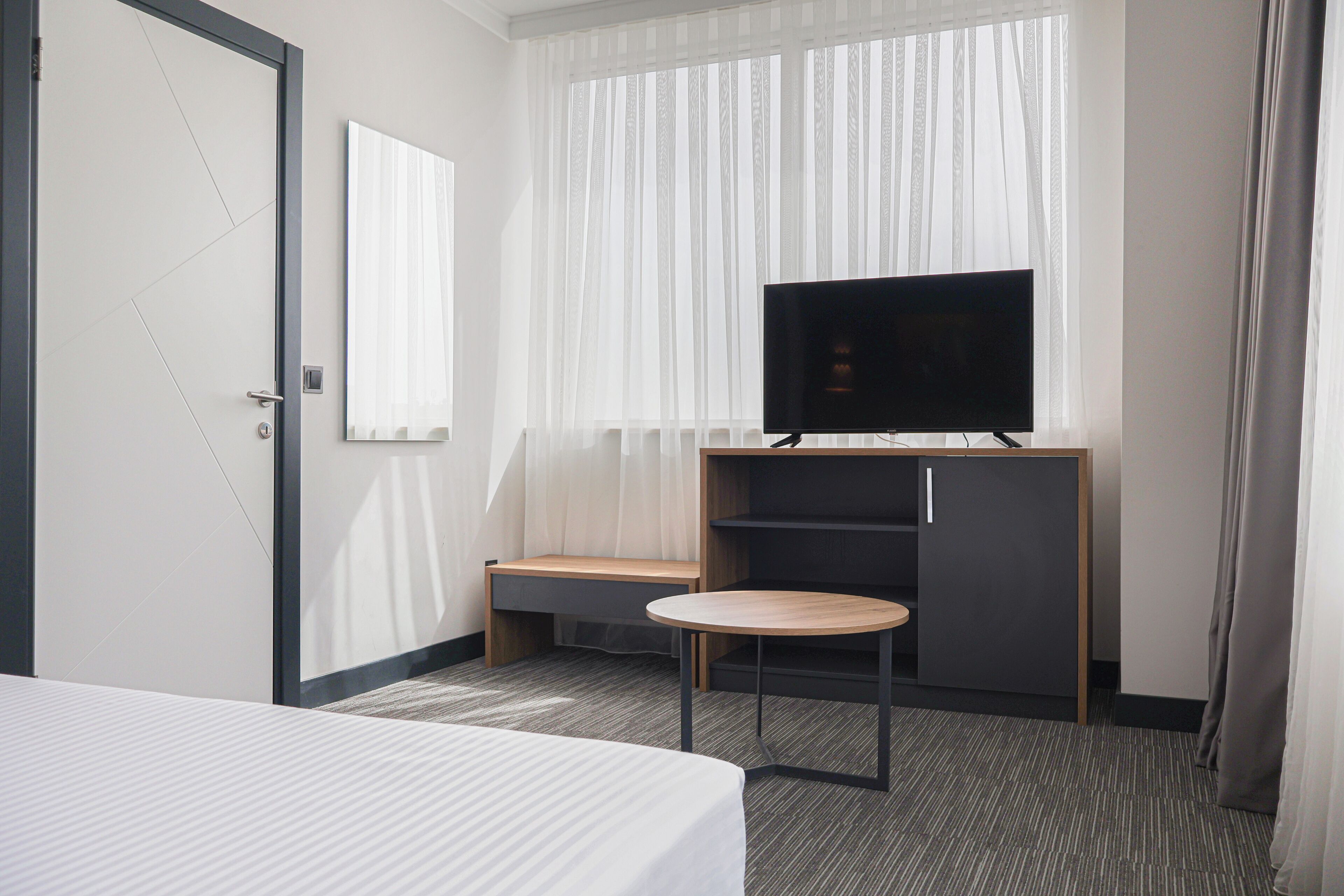 Ein Hotelzimmer mit einem Bett, einem Fernseher auf einem Holzboden, einem kleinen runden Tisch und einem Fenster mit durchscheinenden Vorhängen.