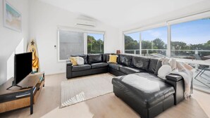 A modern living room with a black leather sectional sofa, a flat-screen TV, and a large window offering a view of greenery. - Seaworthy Sanctuary • Stylish Coastal Escape (Jan Juc)