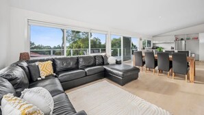 A modern living room with a sectional sofa, a dining area with a wooden table and chairs, and a kitchen with white cabinets and stainless steel appliances. - Seaworthy Sanctuary • Stylish Coastal Escape (Jan Juc)