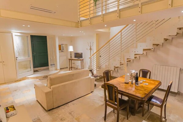 A spacious living area with a sofa, a dining table with chairs, a staircase, and a television.