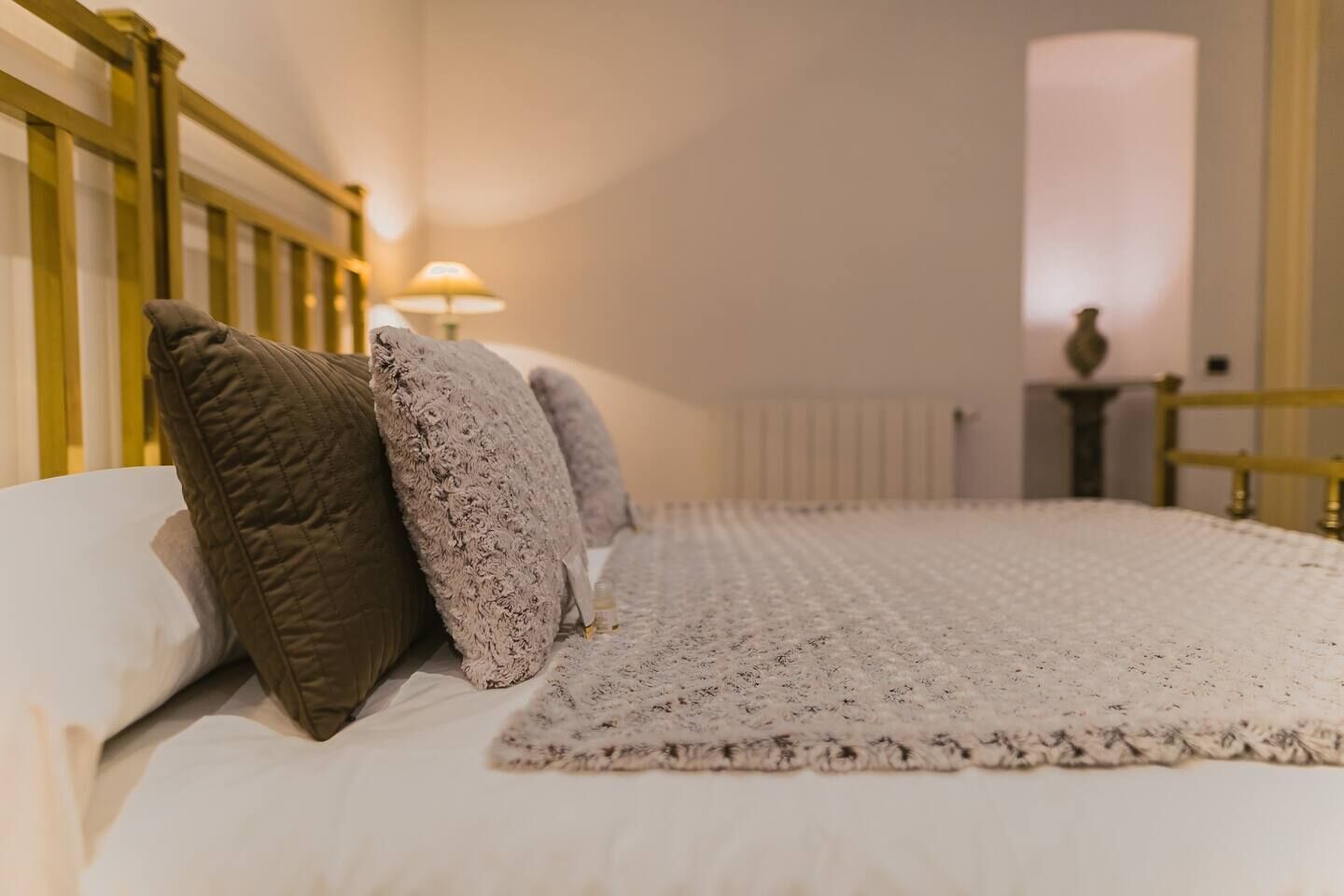 A neatly made bed with a white duvet and a lace bedspread, a wooden headboard, and a lamp in the background.