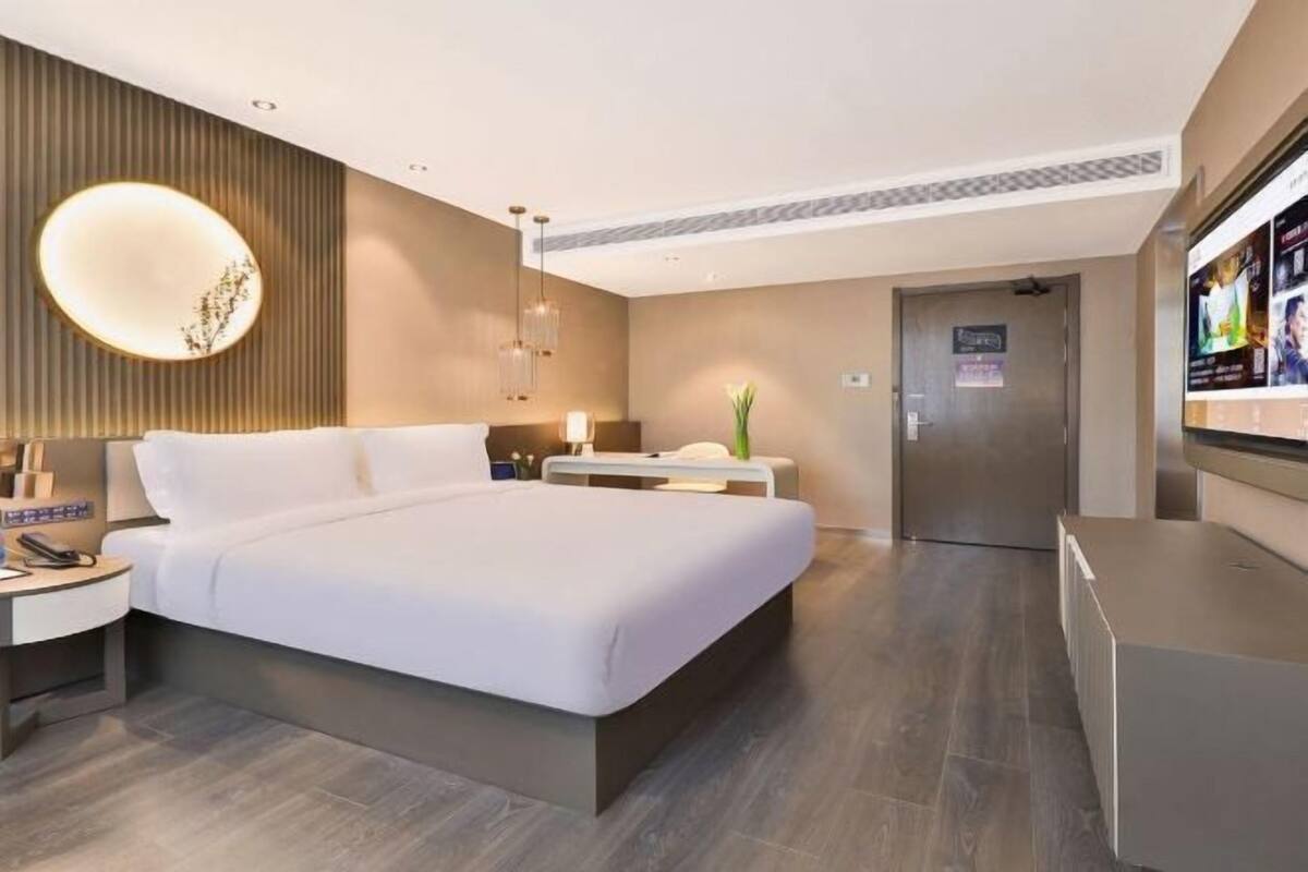 A modern hotel room with a large bed, bedside tables, a desk, a chair, a TV, and a door.
