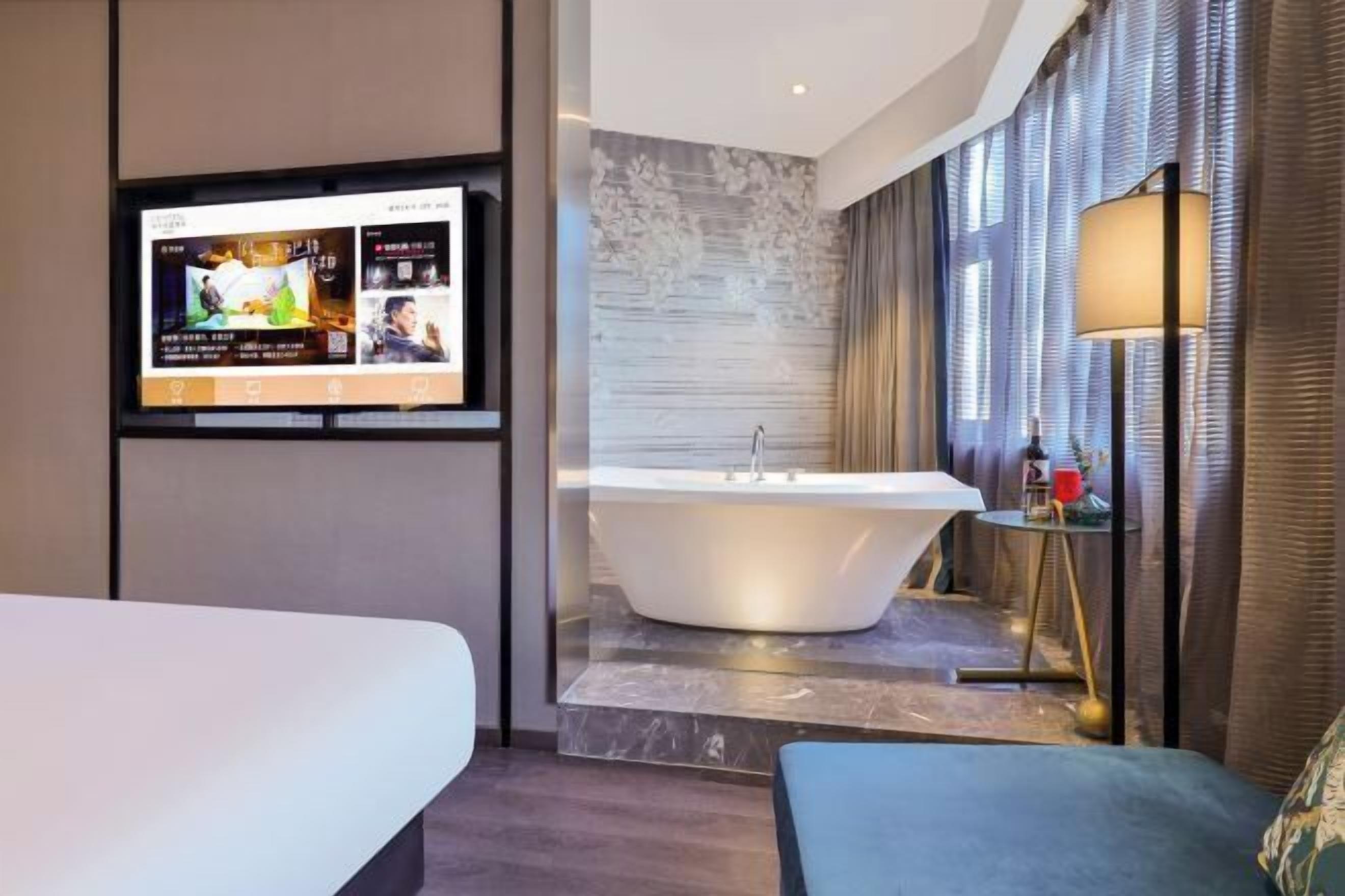 A modern hotel room with a large flat-screen TV mounted on the wall, a freestanding bathtub, and a comfortable seating area with a sofa and armchair.