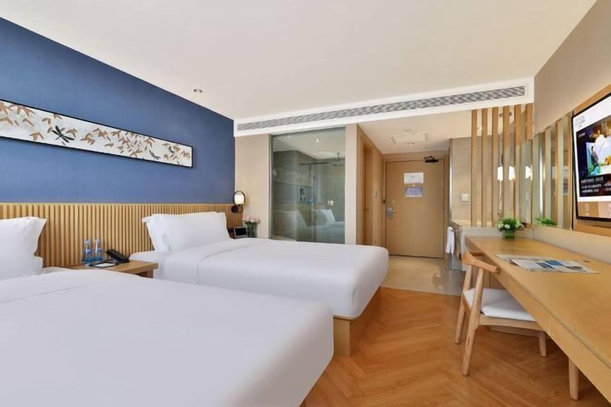 A hotel room with two beds, a desk, a TV, and a bathroom visible through a glass door.