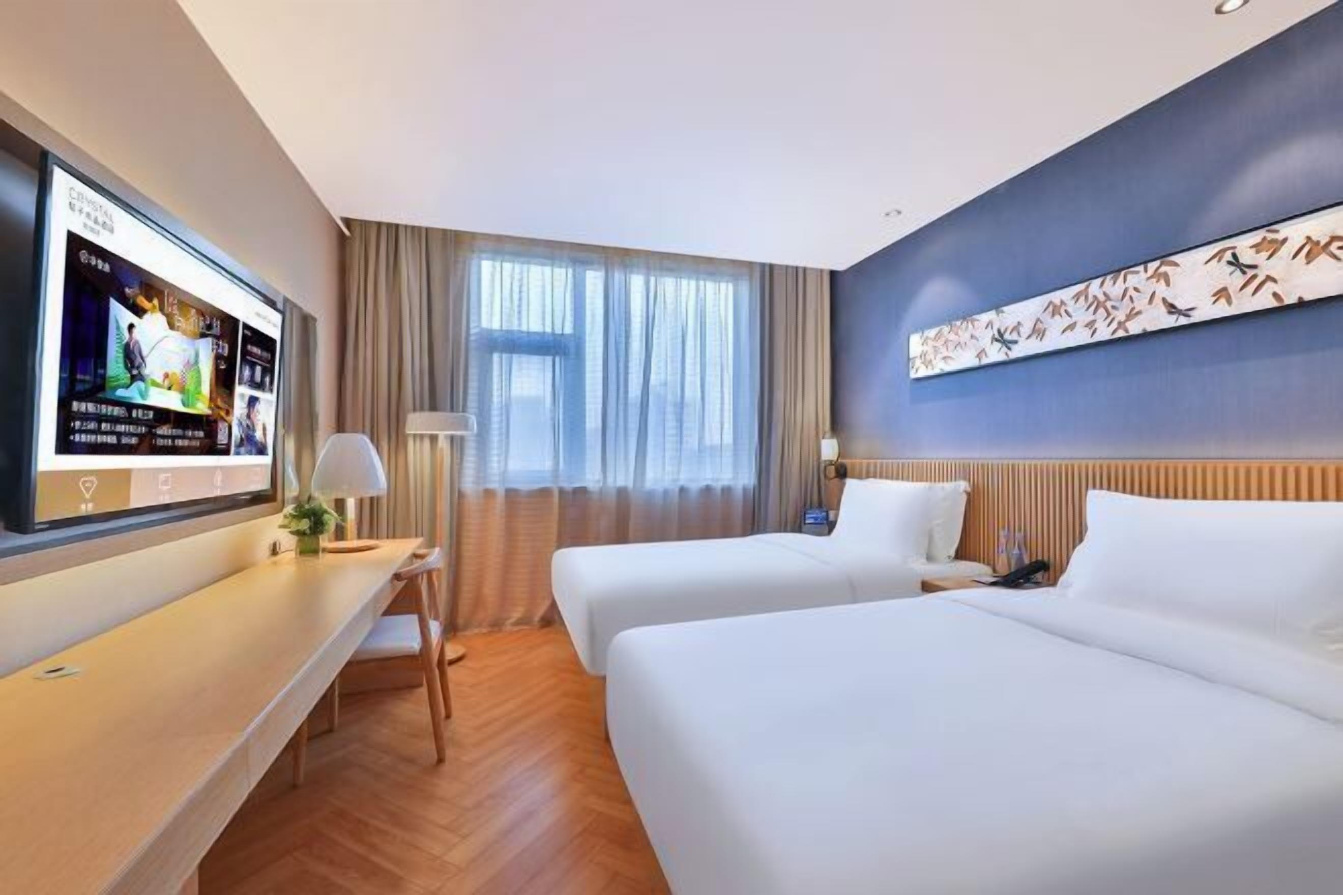Une chambre d’hôtel avec deux lits, un téléviseur à écran plat fixé au mur, un bureau avec une lampe et une grande fenêtre avec des rideaux.