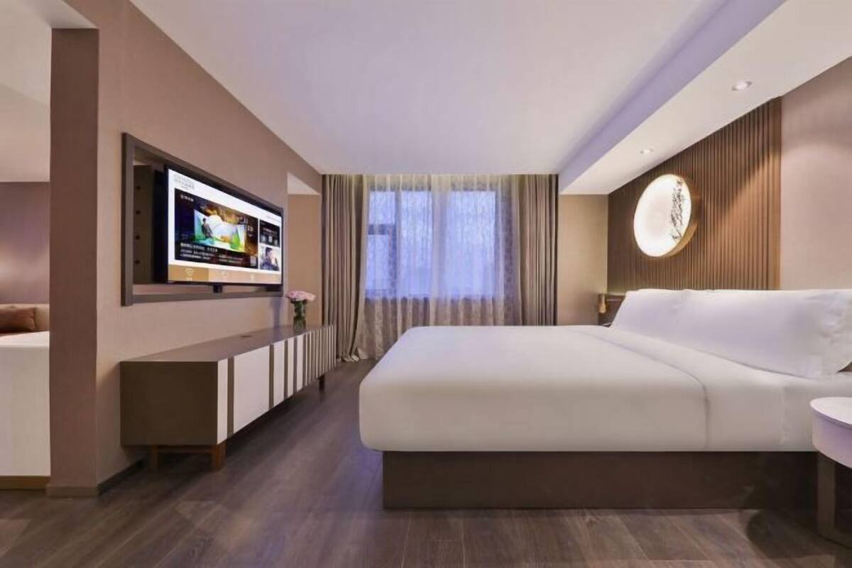 A modern hotel room with a large bed, a TV mounted on the wall, a small side table, and a window with curtains.