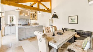 Une cuisine moderne dotée d’un îlot central, de poutres en bois, de luminaires suspendus et d’un coin repas avec un banc et des chaises.