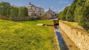 Un pont de pierre enjambe un petit ruisseau, entouré d’une végétation luxuriante et de murs de pierre, et mène à un bâtiment traditionnel en pierre doté de plusieurs cheminées.