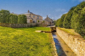 A stone bridge over a narrow stream, surrounded by lush green grass and stone walls, leading to a traditional stone building with multiple chimneys. - Washbrook Cottage (Little Compton)