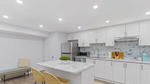 Cocina moderna con armarios blancos, un refrigerador de acero inoxidable, un microondas y un lavamanos. Hay un banco con cojín y dos sillas de mimbre.
