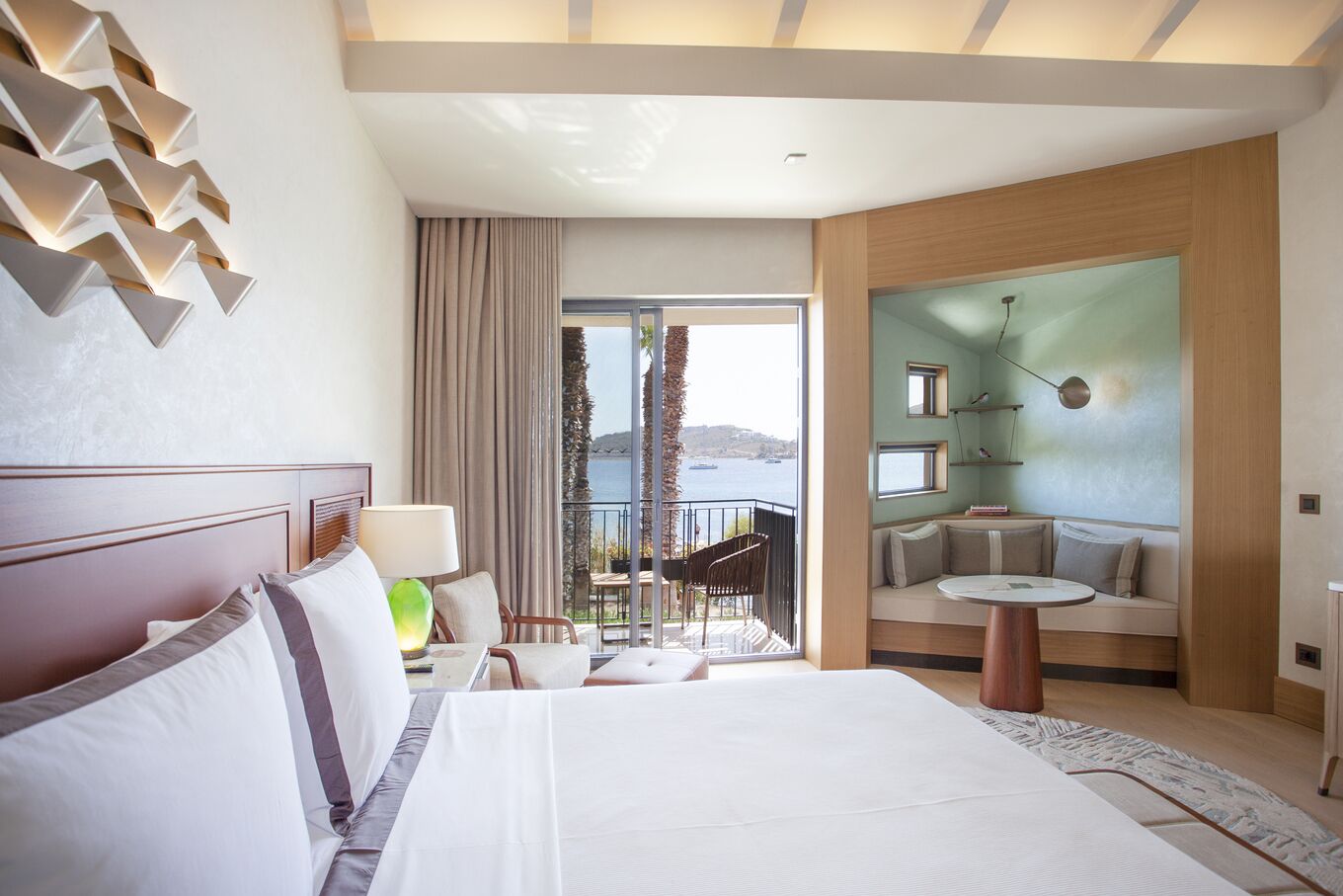 A modern hotel room with a large bed, a balcony with a view of the sea, and a seating area with a table and chairs.