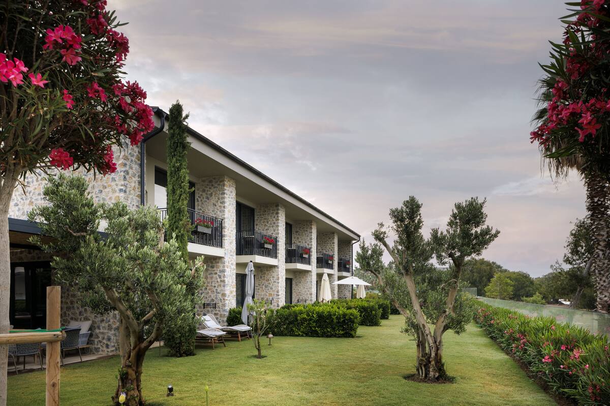 Un hotel con fachada de piedra, varios balcones y un jardín bien cuidado con diversas plantas y una zona de piscina.
