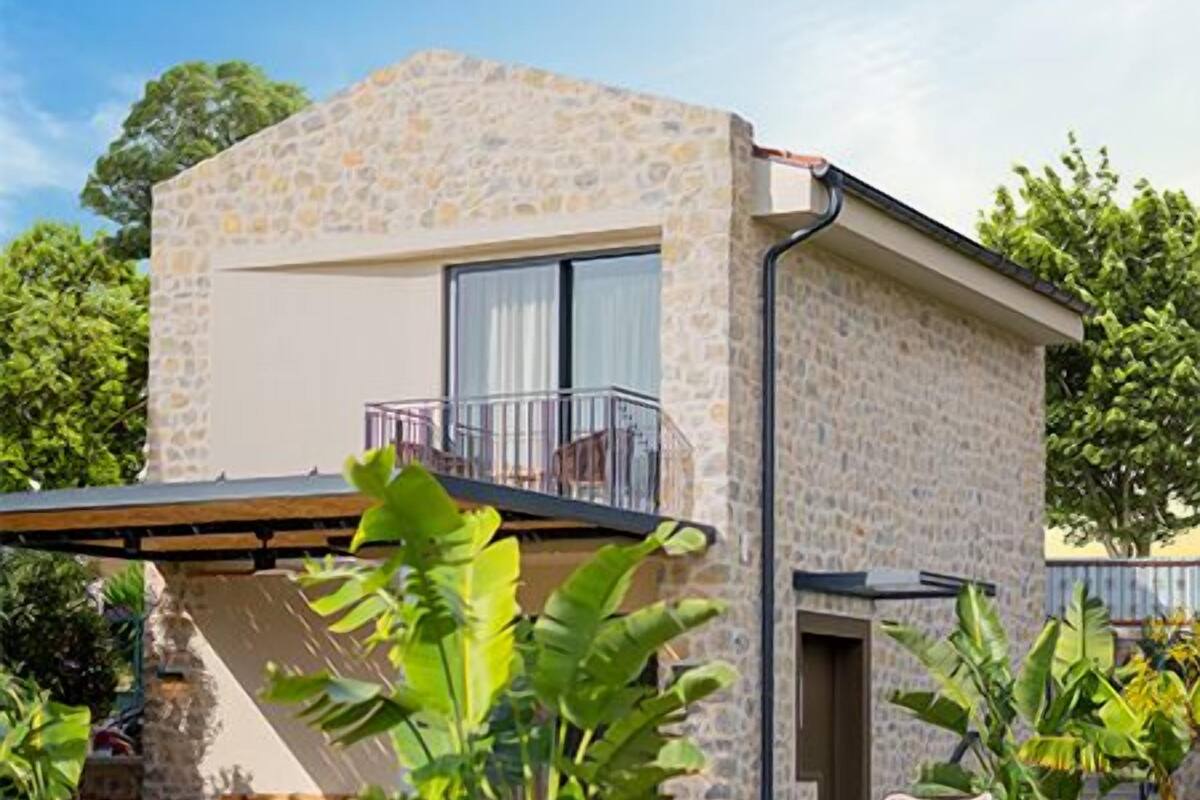 Una casa moderna de dos pisos con fachada de piedra, un balcón con mobiliario exterior y un jardín bien cuidado con diversas plantas y un camino.