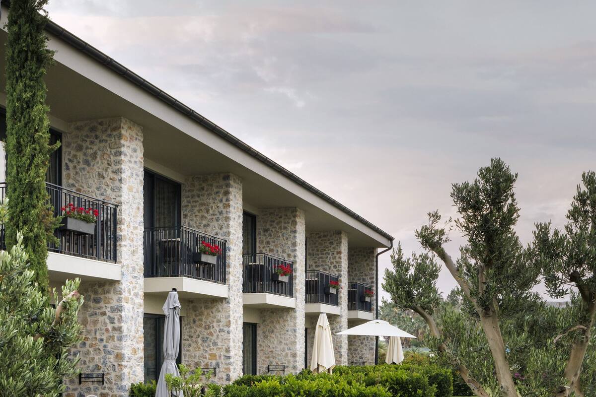 Hotel con fachada de piedra, varios balcones, zona de estar exterior y abundante vegetación.