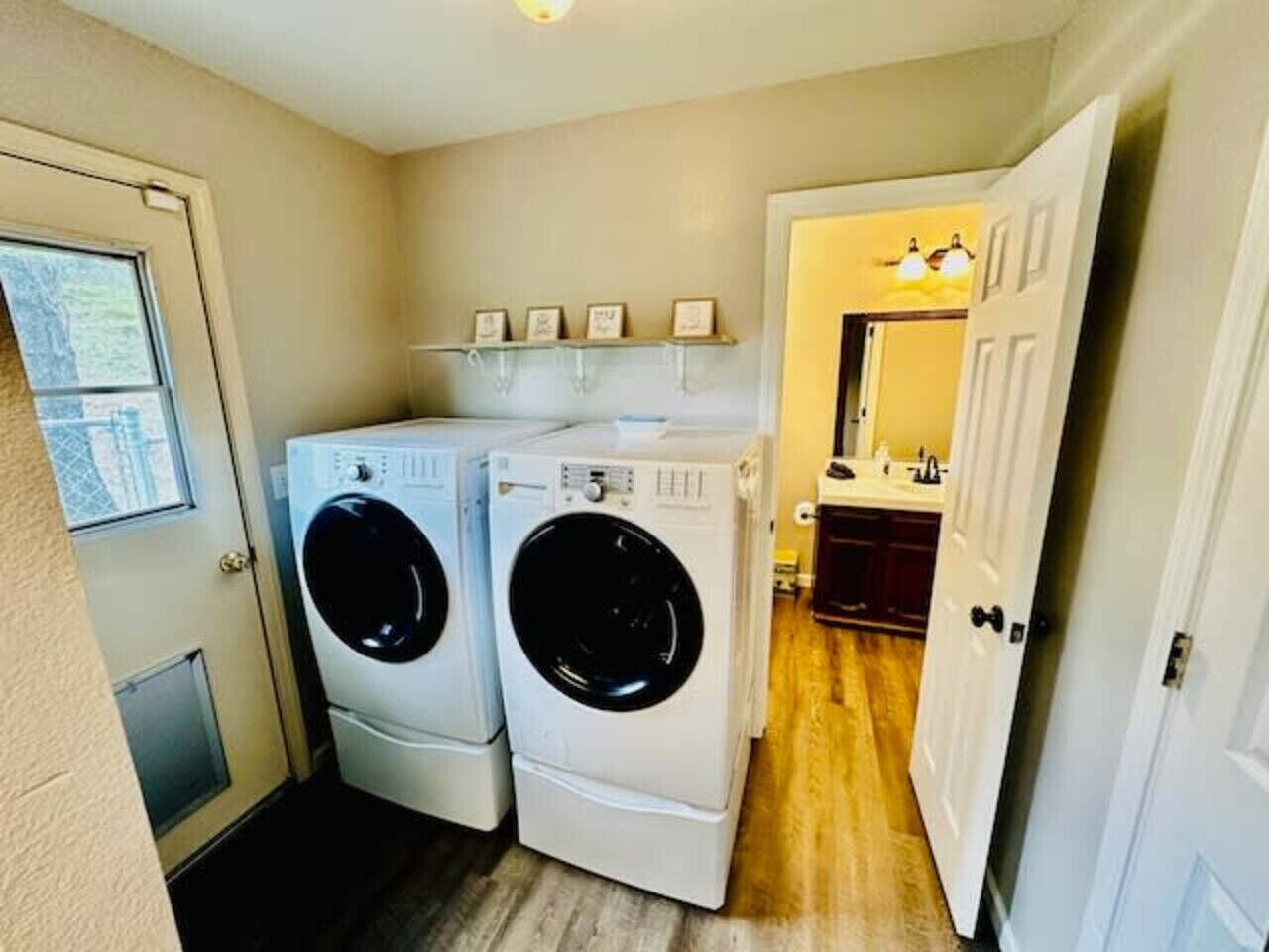 A laundry room with two stacked front-loading washers and dryers, a shelf with framed pictures, and a door leading to a bathroom.