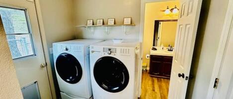 A laundry room with two stacked front-loading washers and dryers, a shelf with framed pictures, and a door leading to a bathroom.