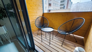En träaltan med två stolar med stomme av metalltråd och ett litet bord, en vit soptunna och en glasdörr som leder in till ett inomhusområde.