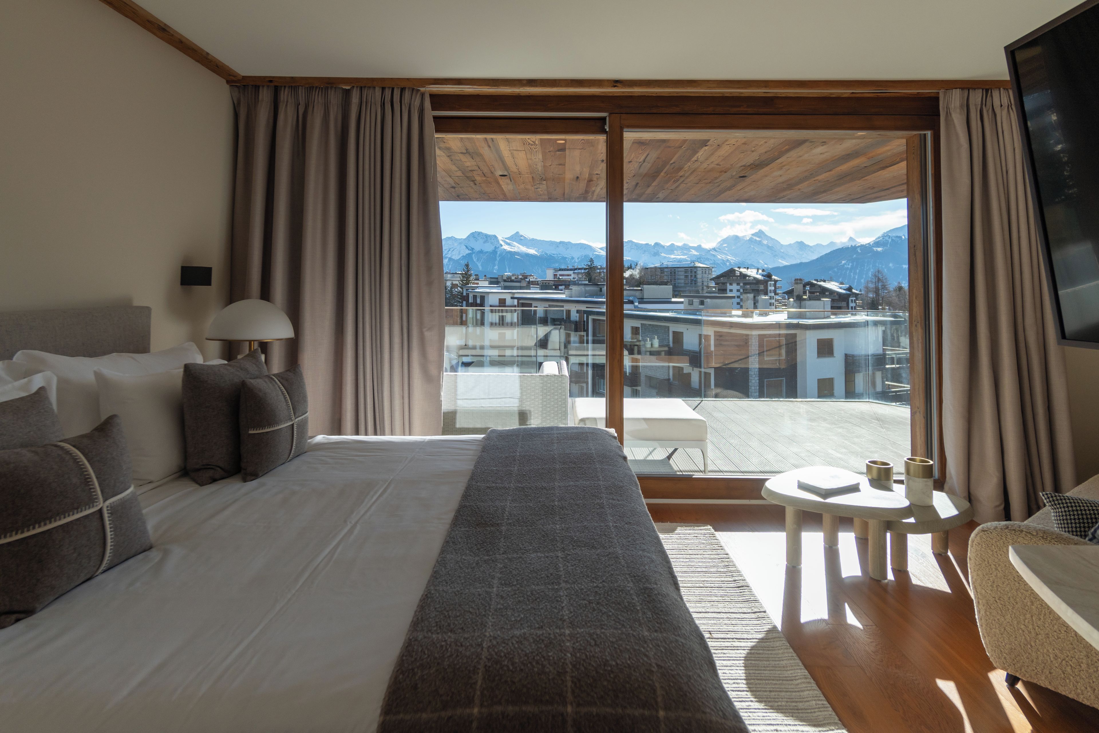 Habitación de hotel con una cama grande, una zona de estar con una mesa y sillas, y vistas a las montañas a través de un ventanal corredero.