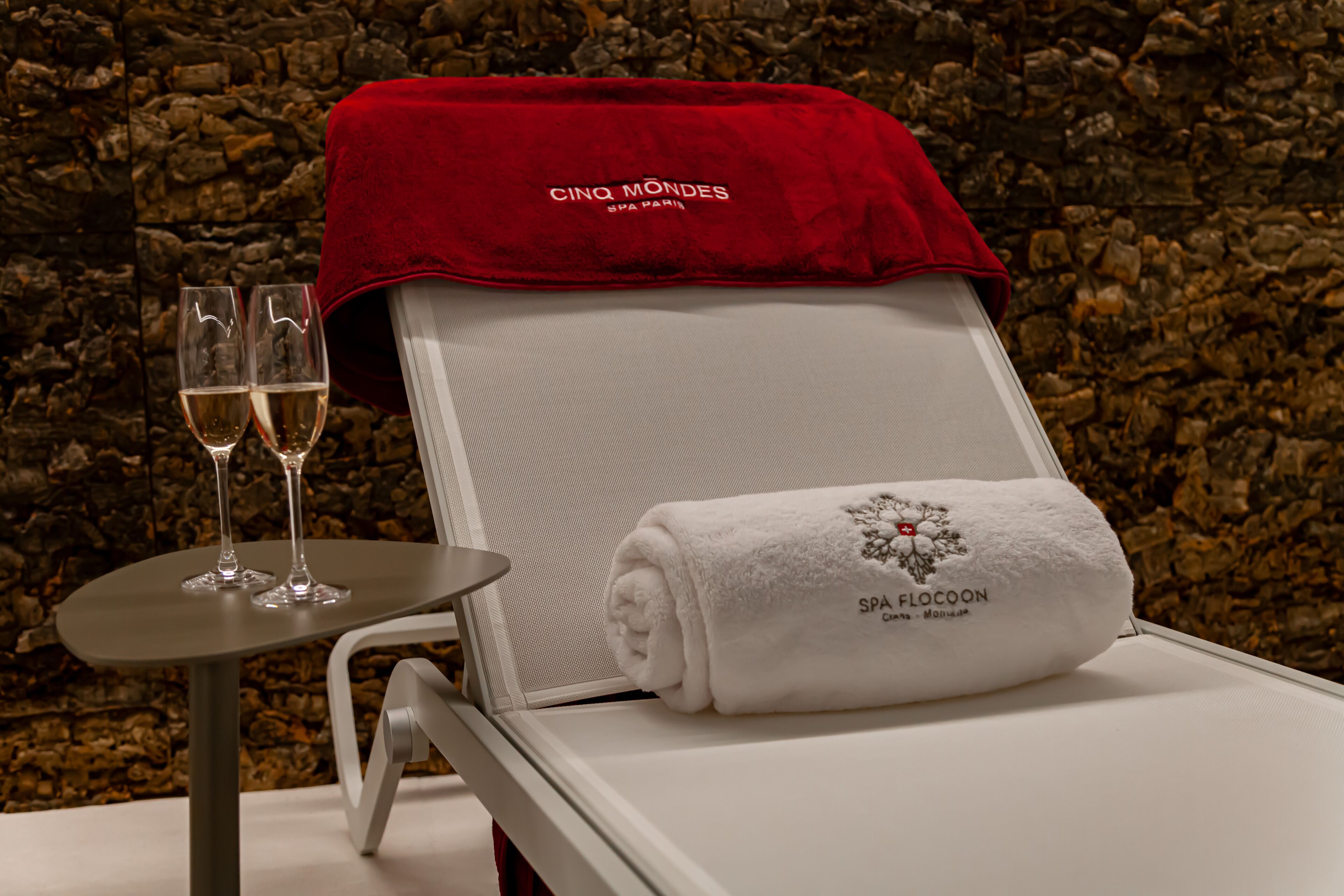 Una tumbona de spa con una toalla blanca y una cubierta roja, dos copas de champán y una mesita redonda.