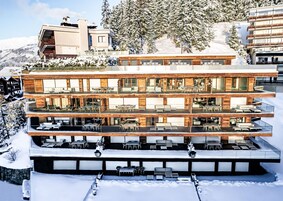 Un hotel de varios pisos con zonas de estar al aire libre en cada nivel, rodeado de árboles nevados y montañas.