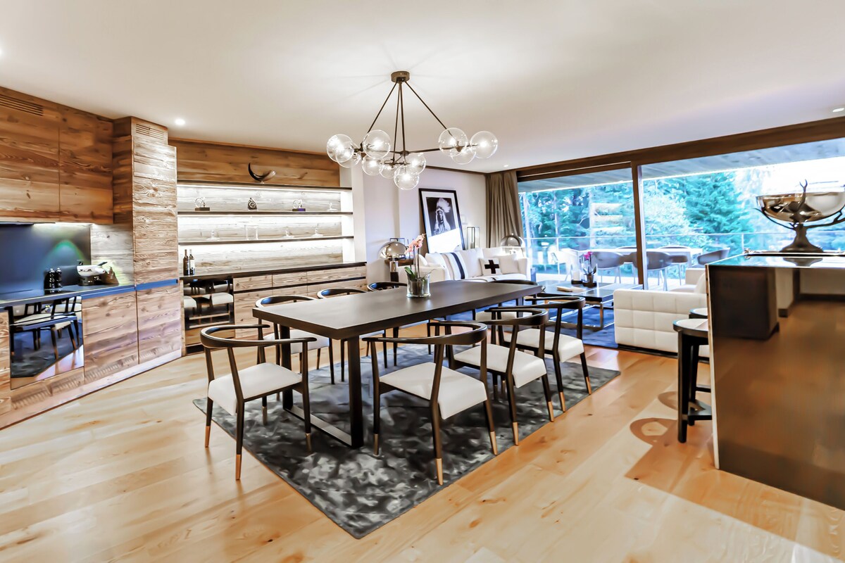 A modern kitchen with wooden accents, a dining area with a chandelier, and a large window offering a view of the outdoors.