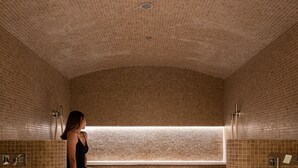 Eine Frau sitzt in einer Spa-Badewanne mit gefliesten Wänden und Boden, einer Sitzbank und einem Fenster mit natürlichem Licht.