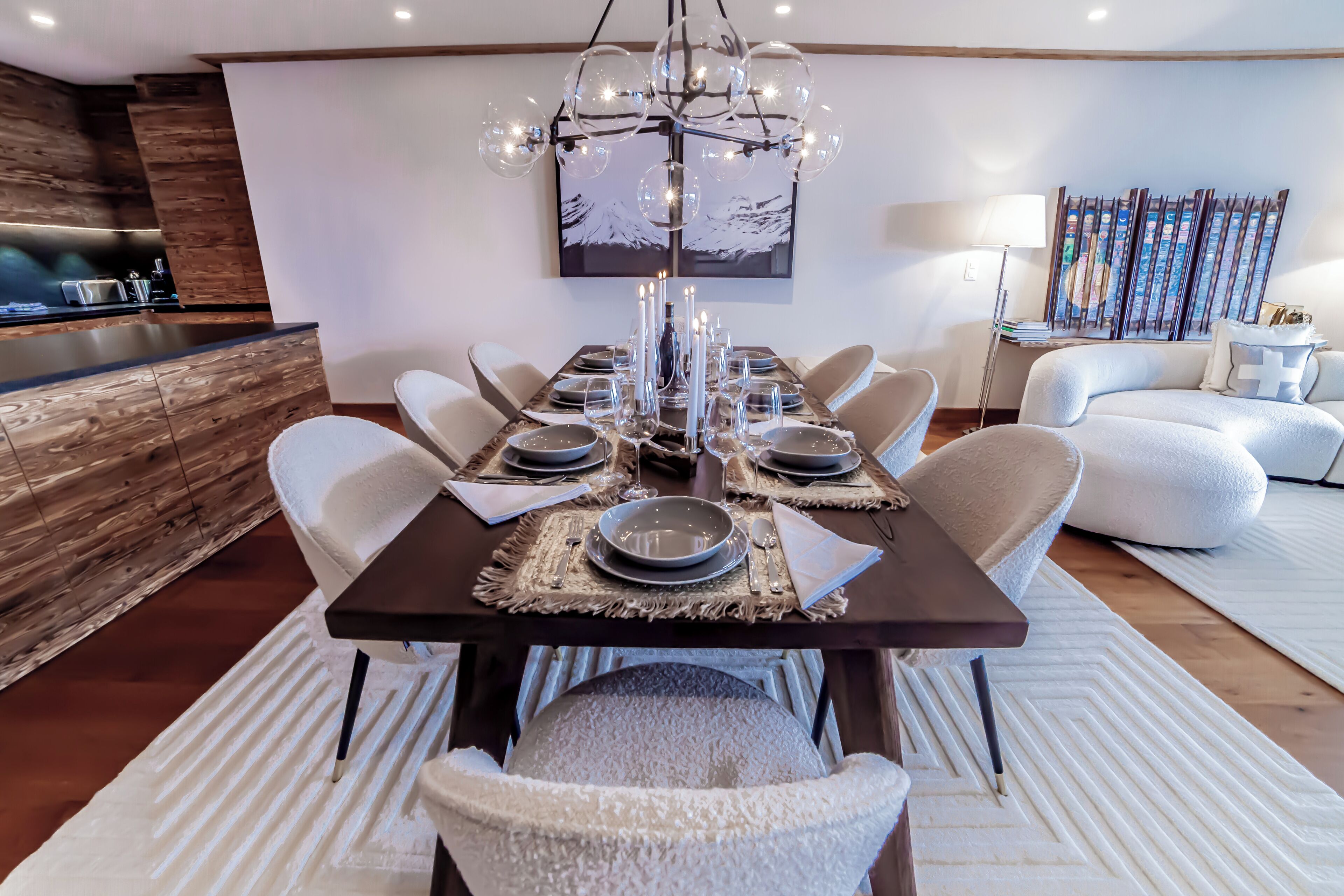 A modern dining area with a long table set for six, surrounded by white cushioned chairs.