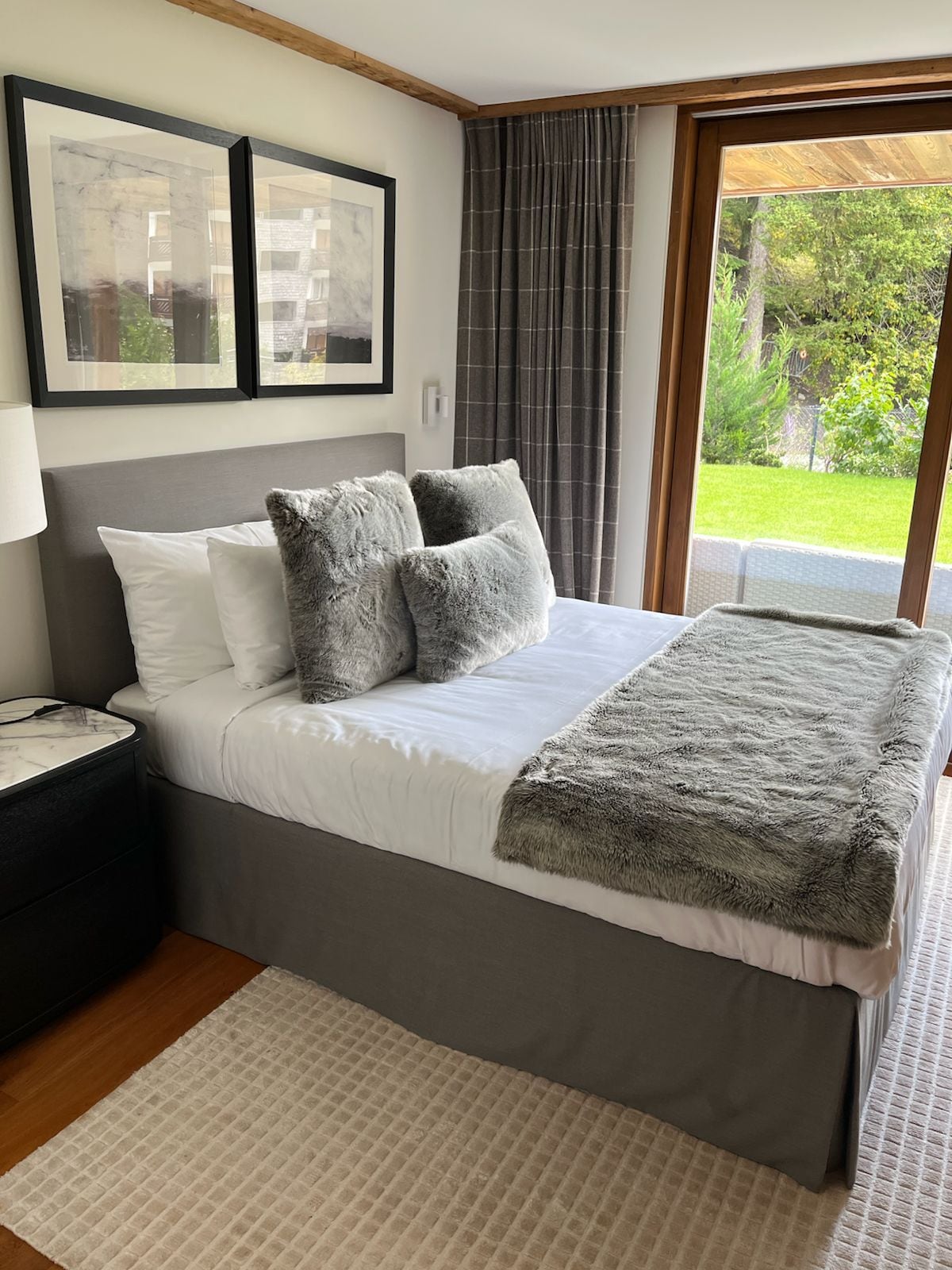 Une chambre à coucher avec un lit, deux tableaux encadrés, une table de chevet, une lampe, une fenêtre avec des rideaux et une vue sur la verdure à l’extérieur.